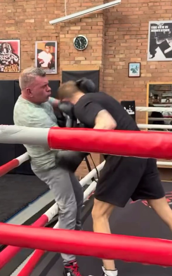Two men sparring in the gym's boxing ring, with brick walls and framed posters behind
